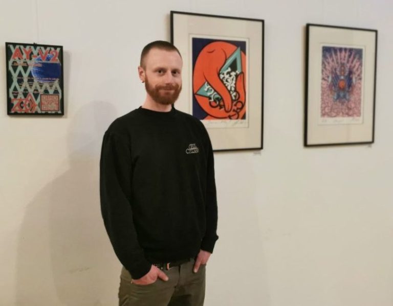 Ich bins Mann steht neben bunten Kunstwerken an einer Wand in einem Ausstellungsraum.